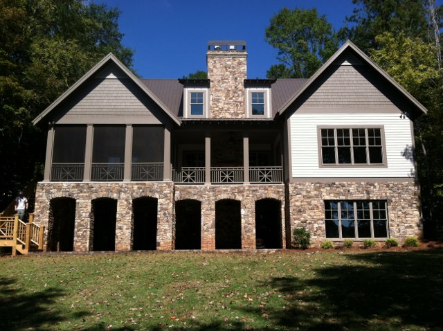 Two-story house with stone lower level, white siding above, gray trim, large windows, porch, arches, stone chimney, grass and trees.
