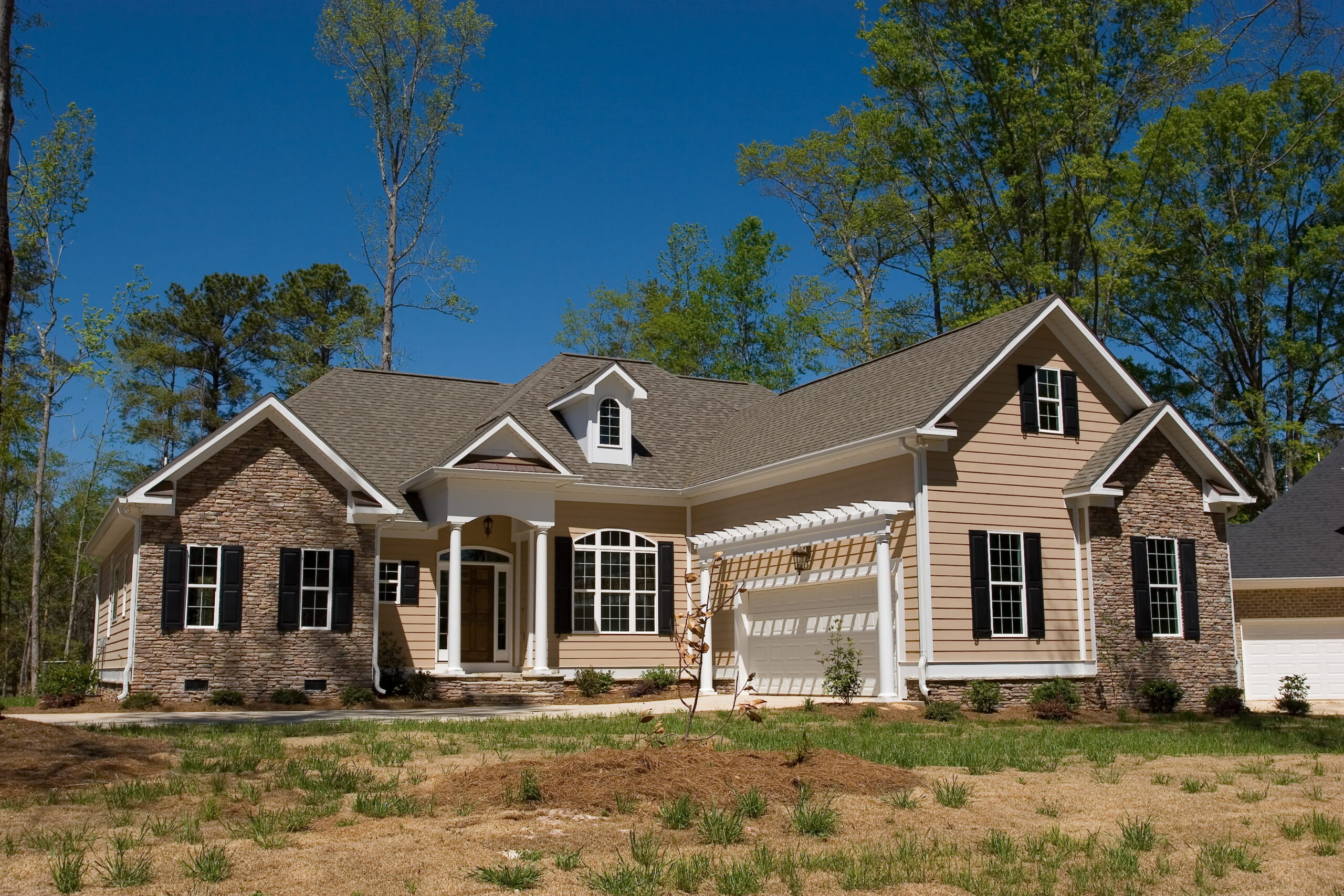 Single-story suburban house with stone and beige siding, white trim, gables, covered entry, pergola, sparse grass, trees, blue sky.