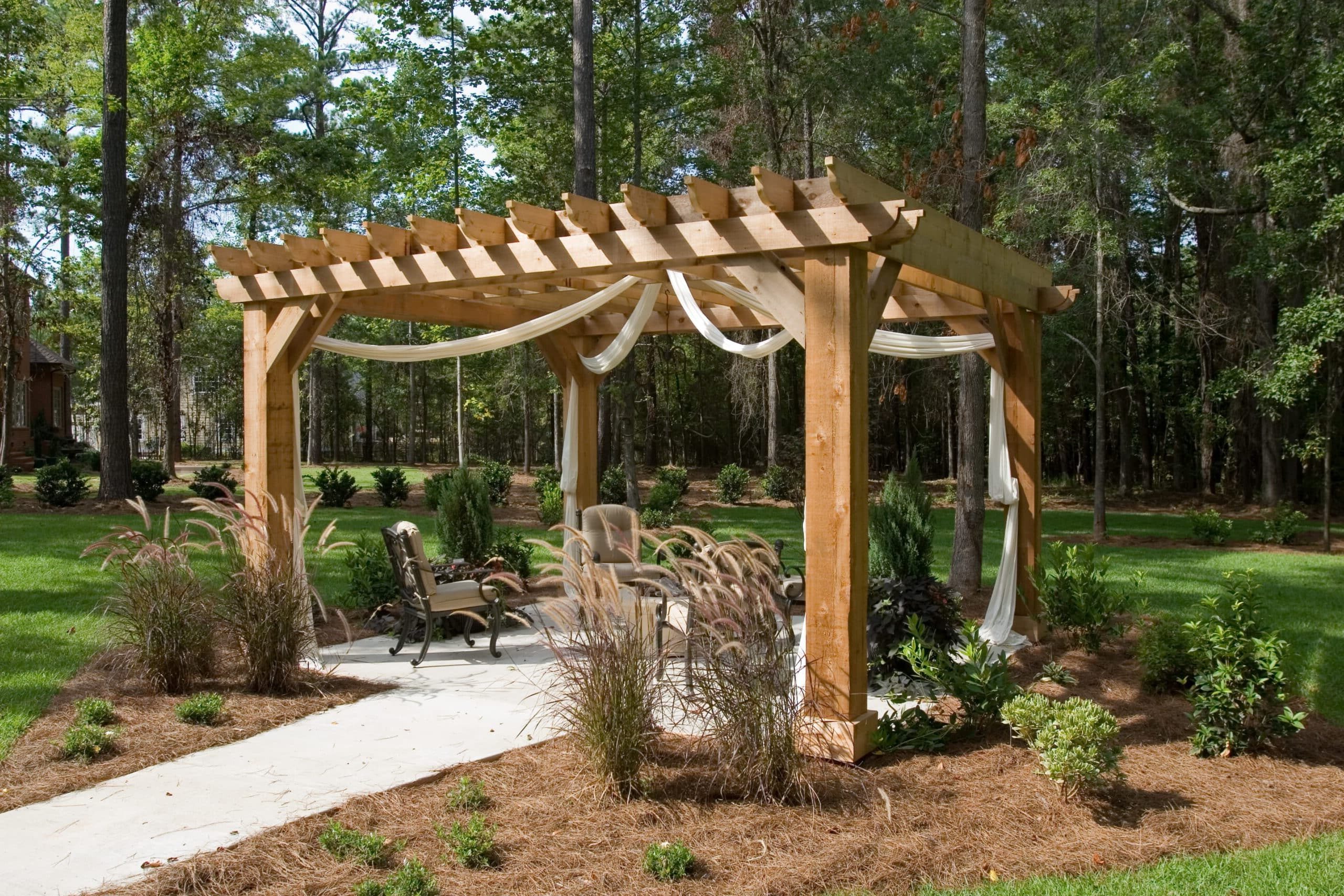 A wooden pergola with white drapes shades a small seating area amid green plants and grasses in a landscaped yard with trees.