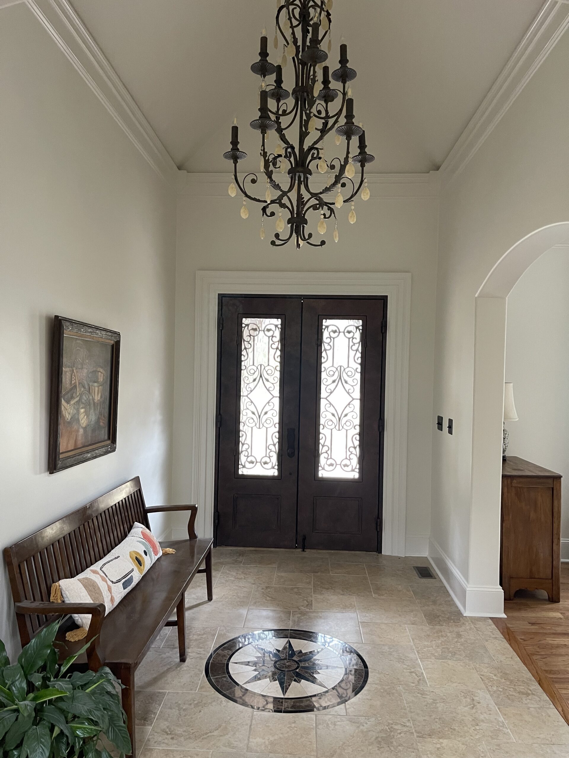 Bright entryway with double wooden doors and glass panels, black chandelier, wood bench with colorful pillows, medallion, light walls.