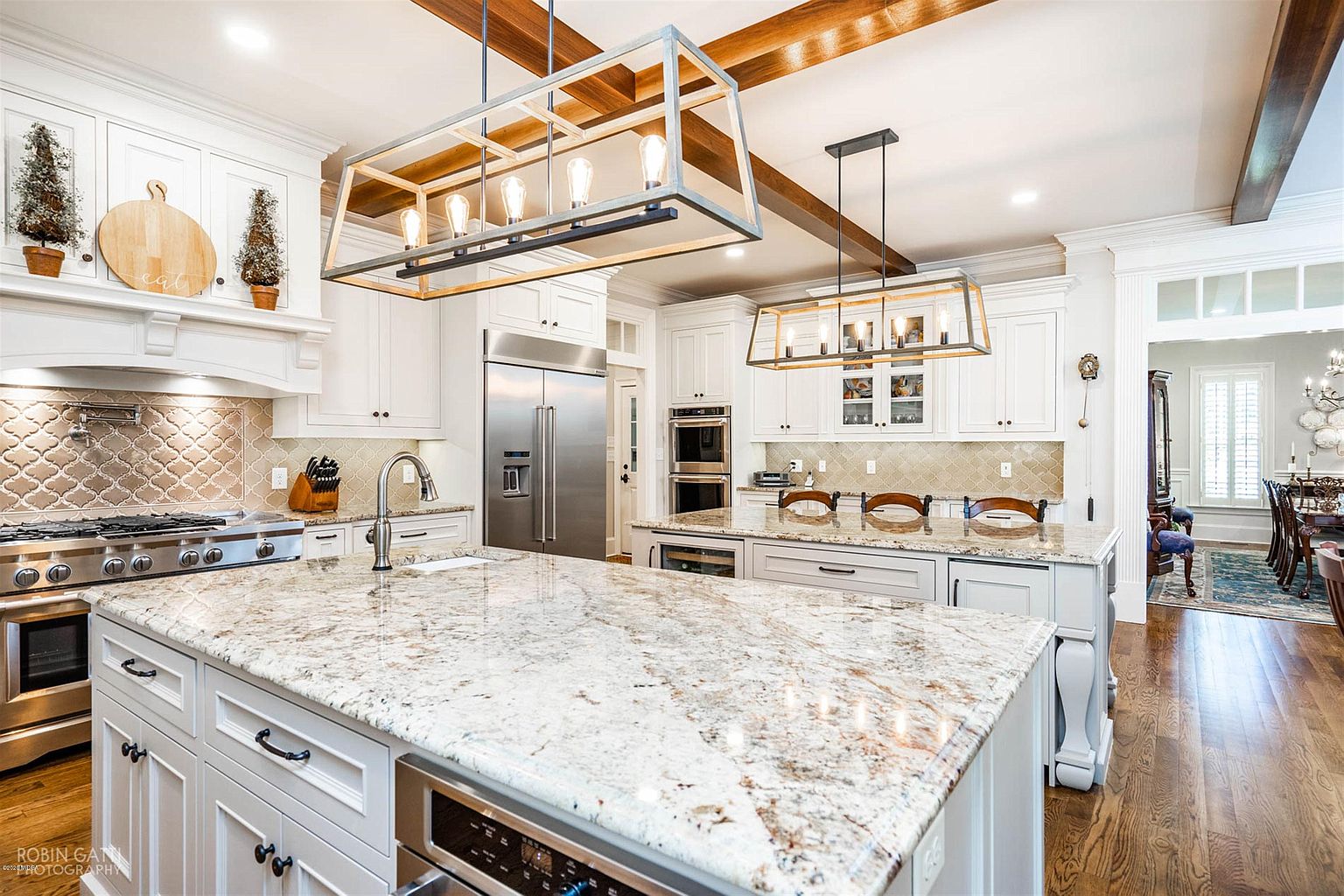 Spacious modern kitchen featuring a large granite island, white cabinets, stainless appliances, double ovens, and exposed beams.